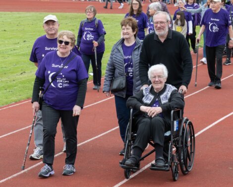 Group of older adults, some wearing purple "Survivor" shirts, participating in a relay event on a track. A woman in a wheelchair is being pushed by a man while others walk with canes.