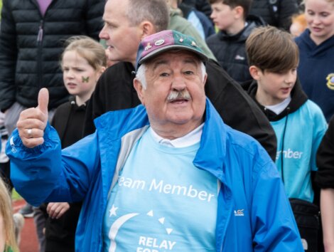 Elderly man in a blue jacket and cap gives a thumbs-up while participating in a community event, wearing a "Team Member" shirt. Several others, including children, are seen in the background.