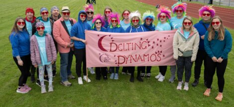 A group of people, some wearing colorful wigs and red heart-shaped glasses, stand on a field holding a pink banner that reads "Delting Dynamos.