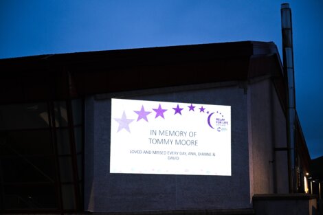 A tribute message displayed on an outdoor screen that reads: "In memory of Tommy Moore. Loved and missed every day, Ann, Dianne & David." The background shows an evening sky.