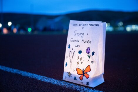 A luminaria bag labeled "Granny + Granda Purdie, Gone but never forgotten" with a butterfly and flower drawings, is placed on a track, illuminated from within at dusk.