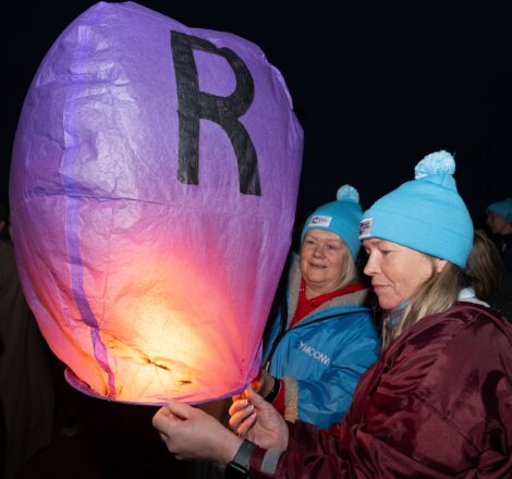 Two people wearing blue beanies light a purple sky lantern with the letter "R" on it during a nighttime event.