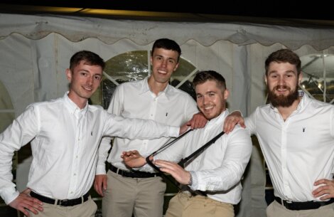 Four men wearing white shirts and beige pants pose together under a tent. One man is playfully tugging on another's suspenders. All appear to be enjoying the moment.