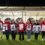 A group of eight people stand in a row holding large signs with numbers and symbols, displaying the total amount raised as £17,418.68. They are in front of a white tent at night.