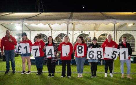 A group of eight people stand in a row holding large signs with numbers and symbols, displaying the total amount raised as £17,418.68. They are in front of a white tent at night.