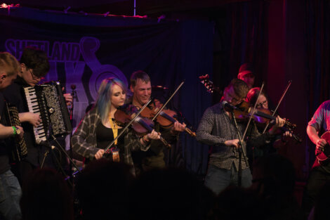 A group of musicians, including violinists and an accordion player, perform on stage at the shetland folk festival.