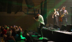 A violinist performs energetically on stage at a concert with a saxophonist and guitarist in the background, in front of an audience.