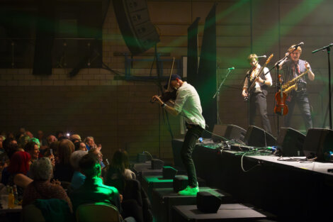 A violinist performs energetically on stage at a concert with a saxophonist and guitarist in the background, in front of an audience.