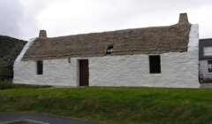 A traditional whitewashed stone cottage with a thatched roof, two chimneys, and a single door, set against a cloudy sky.