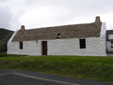 A traditional whitewashed stone cottage with a thatched roof, two chimneys, and a single door, set against a cloudy sky.