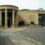 Exterior view of a modern building with large columns and a crest on the sand-colored facade, located beside a street with pedestrians walking by.