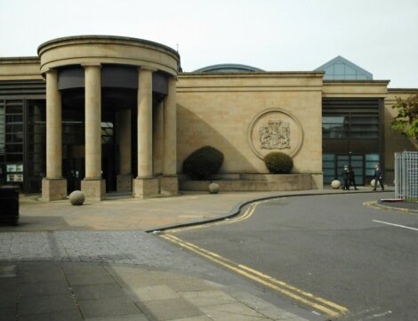 Exterior view of a modern building with large columns and a crest on the sand-colored facade, located beside a street with pedestrians walking by.