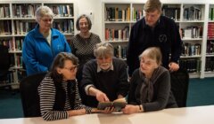 Group of five elderly people, four women and one man, examining an old book together in a library setting.