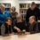 Group of five elderly people, four women and one man, examining an old book together in a library setting.