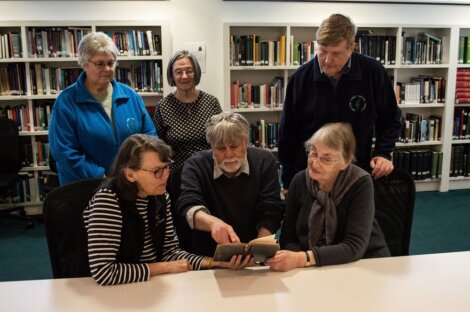 Group of five elderly people, four women and one man, examining an old book together in a library setting.