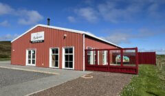 A red and white building labeled "Mackenzies farm shop and cafe" with several glass doors and a fenced outdoor area on a clear, sunny day.