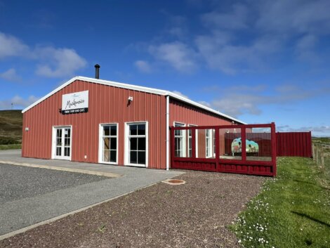 A red and white building labeled "Mackenzies farm shop and cafe" with several glass doors and a fenced outdoor area on a clear, sunny day.