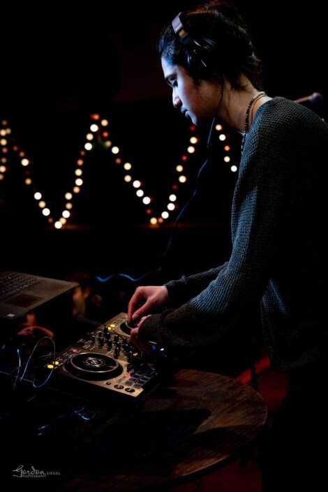 Person wearing headphones operates DJ equipment on a dark stage with string lights in the background.