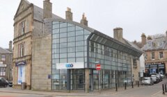 Modern glass facade of a building adjacent to traditional stone structures on a city street, with a "no entry" sign visible.