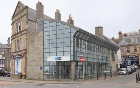 Modern glass facade of a building adjacent to traditional stone structures on a city street, with a "no entry" sign visible.