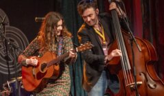 A female guitarist and a male double bassist performing together on stage, both smiling and focused on their instruments.