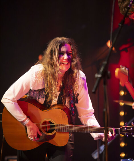 A female guitarist smiling as she plays on stage, lit by ambient warm lighting. she wears a white blouse and a black tie, with her long hair styled loosely.