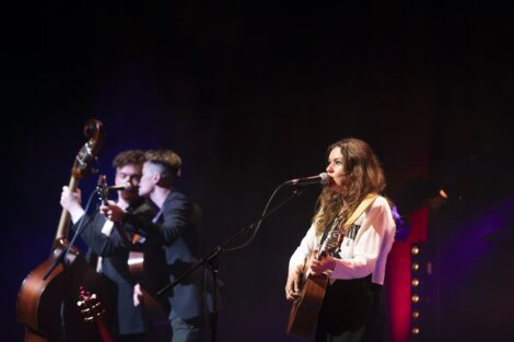 A female singer with long hair performs at a microphone, flanked by two male musicians playing an upright bass and a guitar on a dimly lit stage.