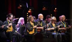 Group of elderly musicians joyfully playing banjos and guitars on stage, highlighted by spotlights in a concert setting.