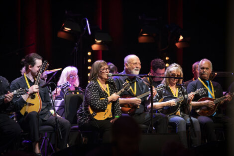 Group of elderly musicians joyfully playing banjos and guitars on stage, highlighted by spotlights in a concert setting.