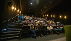 Audience seated in a dimly lit theater with stairs on the left, watching a presentation or a movie.