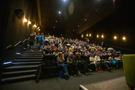 Audience seated in a dimly lit theater with stairs on the left, watching a presentation or a movie.