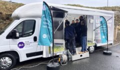 Mobile driving simulation trailer parked with several people standing inside learning about driving tests, affiliated with south lanarkshire college and local councils.