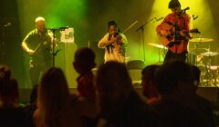 Three musicians perform on stage in front of an audience, featuring a violinist, a guitarist, and another guitarist using an electronic device.