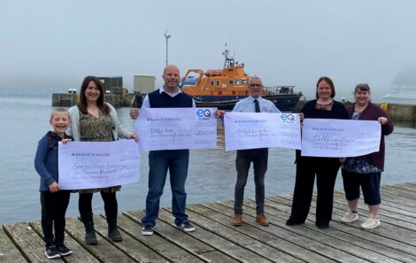 Five people standing on a wooden pier hold large charity checks. A rescue boat is visible on the water behind them. The sky is foggy.