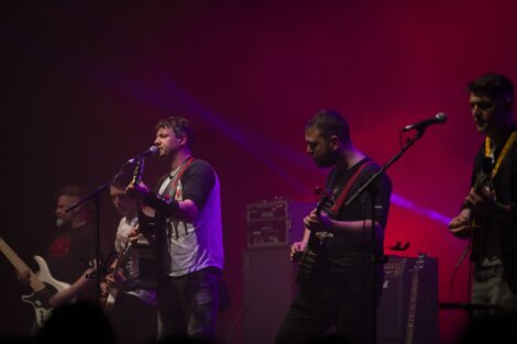 Band performs on stage under purple lighting; lead singer with microphone, others play guitars and bass.