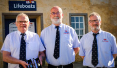 Three men in uniform shirts and ties stand in front of a Lifeboats building, with the man on the left holding two medals.