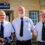 Three men in uniform shirts and ties stand in front of a Lifeboats building, with the man on the left holding two medals.