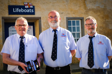 Three men in uniform shirts and ties stand in front of a Lifeboats building, with the man on the left holding two medals.