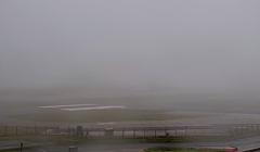 A foggy airfield with limited visibility, featuring runways partially obscured by dense fog, a chain-link fence in the foreground, and faint outlines of buildings in the distance.