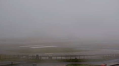 A foggy airfield with limited visibility, featuring runways partially obscured by dense fog, a chain-link fence in the foreground, and faint outlines of buildings in the distance.