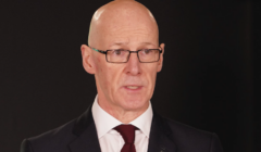 A bald man wearing glasses and a dark suit with a white shirt and burgundy tie speaks in front of a dark background.