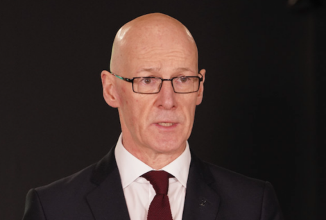 A bald man wearing glasses and a dark suit with a white shirt and burgundy tie speaks in front of a dark background.