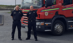 Two firefighters in uniform stand in front of a red fire truck parked in a lot on an overcast day.