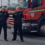 Two firefighters in uniform stand in front of a red fire truck parked in a lot on an overcast day.