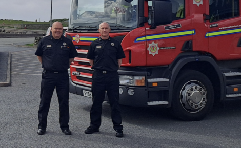 Two firefighters in uniform stand in front of a red fire truck parked in a lot on an overcast day.