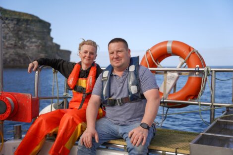 Two people wearing life jackets sit on a boat. The person on the left is in a bright orange jacket, while the person on the right is in a striped shirt. A life preserver is visible in the background.