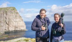 Two individuals stand on a cliffside wearing hoodies and binoculars around their necks, with a rocky coastline and ocean in the background.