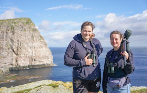 Two individuals stand on a cliffside wearing hoodies and binoculars around their necks, with a rocky coastline and ocean in the background.