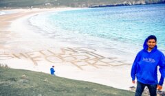 Man in blue hoodie standing on a grassy hill overlooking a beach with "SHETLAND FOSSIL FREE" written in the sand, with a child and cliffs in the background.