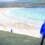 Man in blue hoodie standing on a grassy hill overlooking a beach with "SHETLAND FOSSIL FREE" written in the sand, with a child and cliffs in the background.